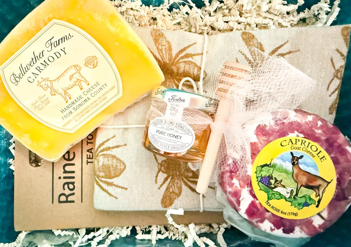 Cheese of the Month Club box featuring two artisan cheeses, a jar of honey, and a wooden honey dipper on shredded paper.